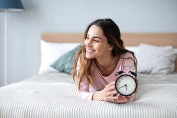 Smiling young woman lying awake in bed with alarm clock, good morning.