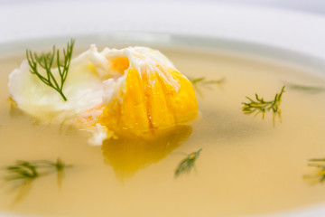 poached egg and dill sprigs in a transparent chicken broth in a white plate close-up