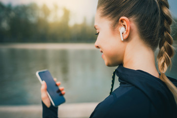 Young smiling girl making sport and running in the park using her phone to listen the music with wireless headphones on sunset in the city watching the screen - obrazy, fototapety, plakaty