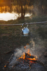 Teapot with tea hangs over the fire on the riverbank sunset