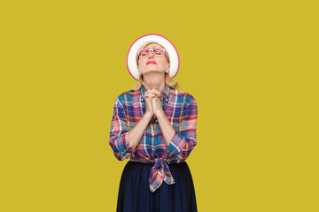 Oh my God please help me. Portrait of hopeful modern stylish mature woman in casual style with hat, eyeglasses standing with closed eyes and praying. indoor studio shot isolated on yellow background.