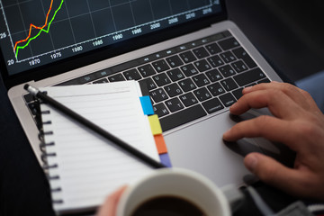 Close-up of man hands, working on touchpad of laptop and holding mug of coffee, near notebook with black pencil.