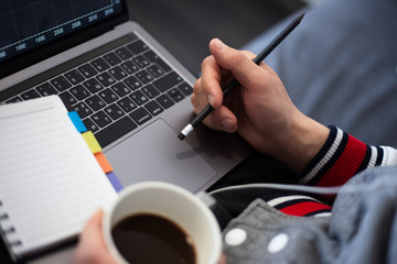 Close-up of man hands, holding cup of coffee and pencil over notebook and keyboard of laptop.