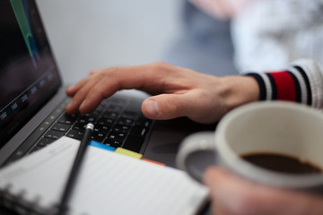 Close-up of man hands, typing on keyboard of laptop and holding mug of coffee, near notebook with black pencil.