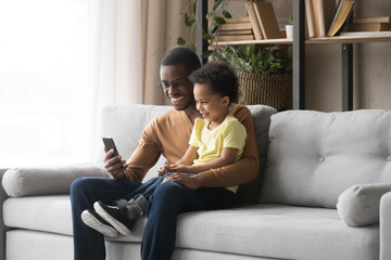 African father son sitting on couch having fun use smartphone