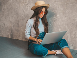 Portrait of creative young smiling woman in sunglasses. Beautiful girl sitting on the floor near gray wall. Model using notebook. Female dressed in hipster clothes and hat.