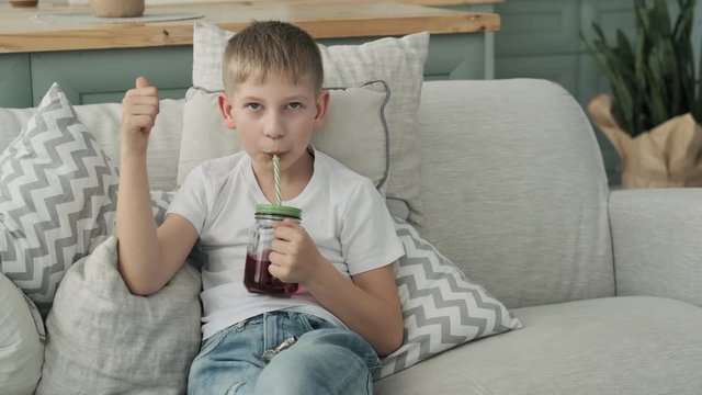 Boy drinking fresh juice sitting at sofa with pillows. Happy boy in white shirt and blue jeans