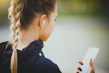 Young smiling girl making sport and running in the park using her phone to listen the music with wireless headphones on sunset in the city watching the screen - obrazy, fototapety, plakaty