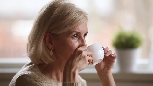 Calm happy old woman sitting alone holding cup enjoying coffee