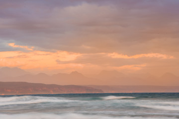 Dreamy blurred waves under an orange and purple sunset sky in Plettenberg Bay, with mountains in the distance. Garden Route, Western Cape, South Africa