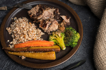 a healthy, tasty meal made of natural ingredients: roasted meat with plums, roasted carrots and groats with the addition of onion served on a rustic plate, next to a fork and knife finished with wood.