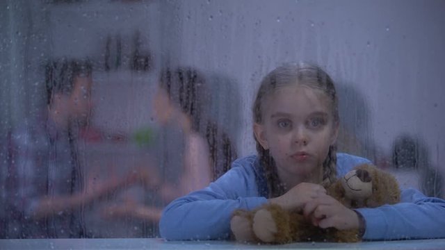 Girl hugging teddy bear on rainy day, father choking mother on background