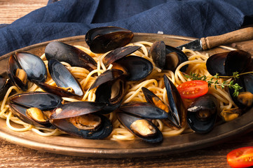 delicious mussels in shells cooked in sauce with pasta and tomatoes on golden metal plate with blue cloth on wooden background