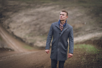 masculine attractive young male in the park or field or track. handsome man in grey coat and blue scarf near the road. affective spring portrait. walking man on the dusty road behind background