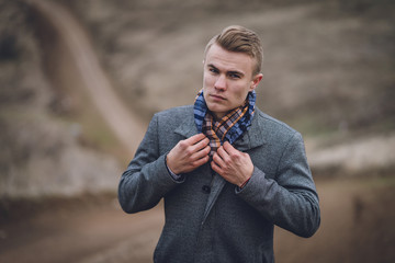 handsome young male man in grey coat and blue scarf near the road track. affective spring portrait. walking man on the dusty road behind background. man straightens scarf looking at camera