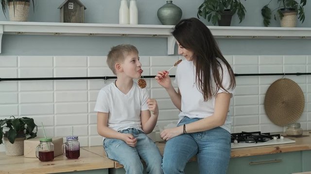 Young mother and son having fun with candy at kitchen. Happy mom spending time with son sitting on table in slow motion