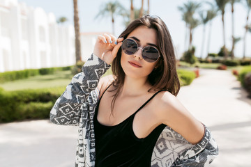 Close-up portrait of trendy girl wearing sunglasses and stylish shirt, posing in the park and enjoying southern sun in vacation. Amazing young woman with dark straight hair standing outside.
