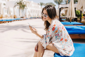 Portrait of brunette girl with straight hair texting message while resting on summer resort. Lovely lady in attire with floral print and sunglasses sitting on daybed under tropical sun and smiling