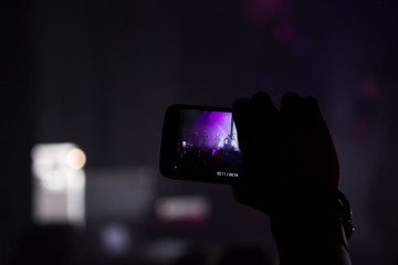 Man hand with a smartphone records a live concert of the group consisting of four cellists and a drummer. The scene is lit with purple spotlights