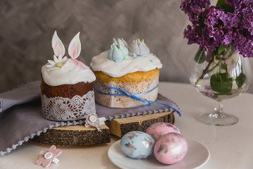 Obraz na płótnie Canvas Easter Cakes on wooden decorated with rabbit ears, eggs on whith plate on foreground, lilac on background. - Traditional Kulich, Paska Easter Bread