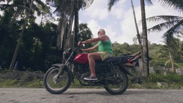 Mature man traveling on motorbike at village road on tropical palm trees landscape. Senior man riding motorcycle on countryside road at summer vacation.