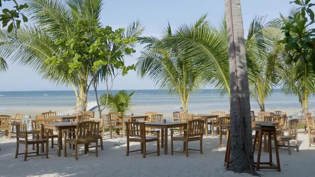 scenic lcoked wide shot of a beautiful beach side restaurant with ocean view, rustic contemporary wooden tables and chairs with shading palm trees, blue sunny day, no people, copy space