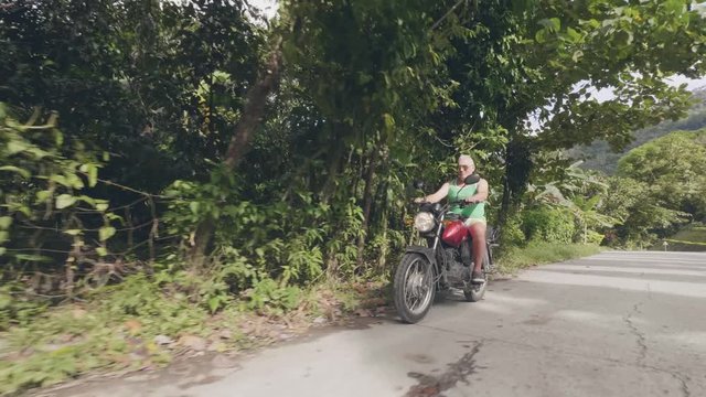 Senior man riding motorcycle on road on tropical village landscape. Mature man driving motorbike while moto trip at summer trip. Motorcycle lifestyle.
