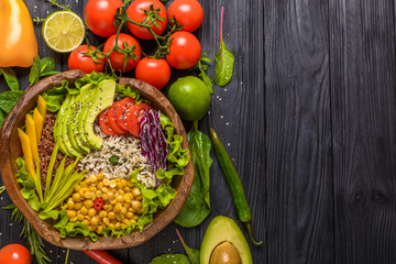 Buddha bowl with chickpea, avocado, wild rice, quinoa seeds, bell pepper, tomatoes, greens, cabbage, lettuce on old black wooden table. Top view with copy space. Healthy vegan food.