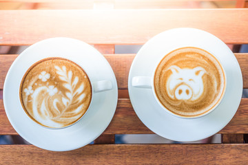 Two cups of coffee with beautiful leaf and pig latte art and sun flare lighting on a wooden table in a coffee shop in the morning.