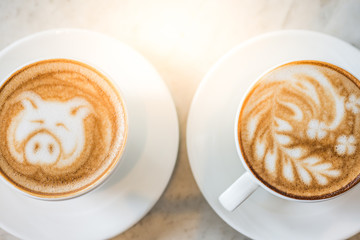 Two cups of coffee with beautiful leaf and pig latte art and sun flare lighting on a marble table in a coffee shop in the morning.