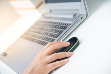 Fototapeta premium Closeup woman's hands clicking mouse and working on a laptop computer on a white desk with sun flare lighting effect .