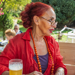 Attractive senior woman in red clothes and round eyeglasses sits on the terrace of a summer park and drinks a beer.