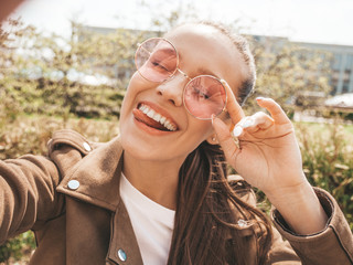 Portrait of beautiful smiling brunette girl in summer hipster jacket and jeans clothes. Model taking selfie on smartphone. Woman making photos in warm sunny day in the street. In sunglasses