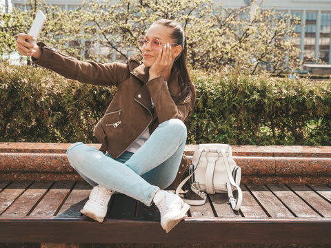 Portrait of beautiful smiling brunette girl in summer hipster jacket and jeans.Model taking selfie on smartphone.Woman making photos in warm sunny day in the street. Sitting on the bench in sunglasses