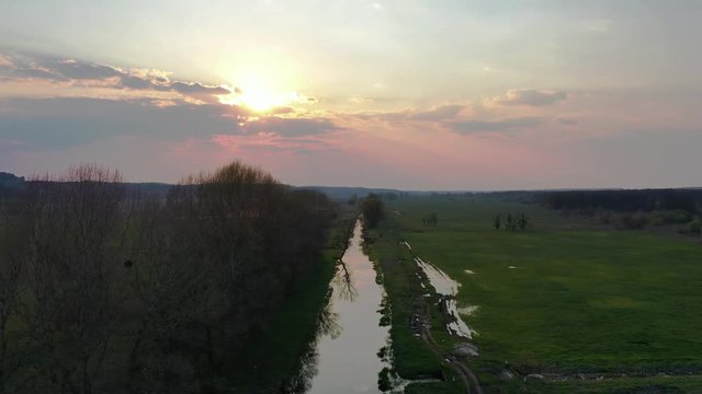 Aerial Flying along river shore. Beautiful early spring landscape with dried canes and trees near water and pine forest, evening, sunset, twilight.