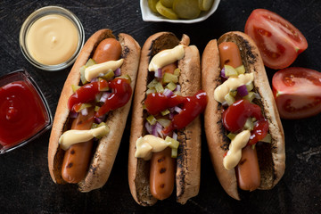 Top view of three hot-dogs with grilled sausages, chopped pickles, red onion and dipping sauces