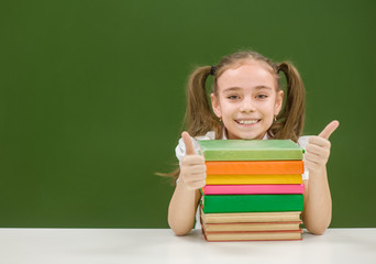 Happy schoolgirl with pile books near empty green chalkboard showing thumbs up. Space for text