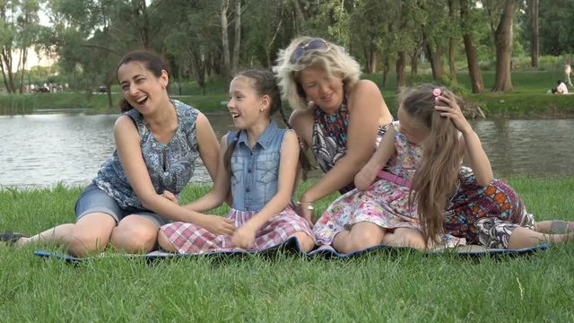 Happy same-sex family: two mothers and two little daughters play and have fun sitting on the grass in the Park at sunset on a summer day against the river. Family vacation in nature. 4K