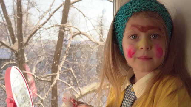 Little cute girl in a mustard jacket and a blue bandage puts makeup looking in a pink mirror, she sits on the windowsill, outside the window is a winter landscape. A child prepare for a carnival.