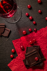 Pieces of black chocolate, cranberries and a glass of red wine on red decorative paper on a black background. For text. top view