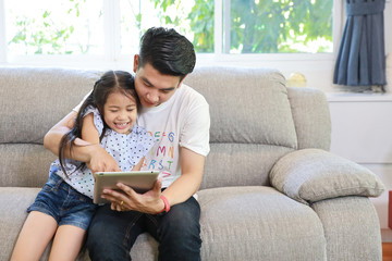 happy asian dad teaching his daughter to use or play tablet on grey sofa in living room with laughing and smiling face (lifestyle with technology concept)