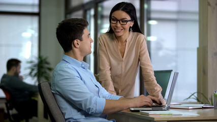 Smiling business lady looking at male colleague sitting laptop, communication