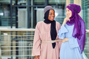 Muslim african fashionably dressed woman talking to her caucasian girlfriend in hijab and blue long dress standing in modern interior