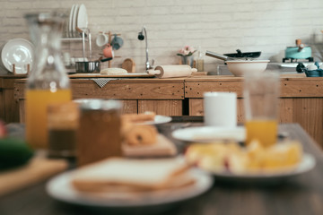 blurred view of healthy meal on kitchen table fresh orange juice toast bread and jam in morning home. wooden cooking place interior in background with handmade dough un bake croissant by rolling pin.