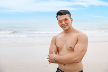 portrait of young asian handsome and sexy man in black swimming suit and sun glasses with smiling face standing on sandy beach with blue sky and left copy space