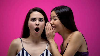 Asian woman whispering secrets to shocked female friend, interesting gossip