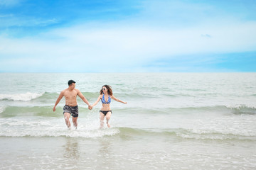 Fototapeta na wymiar cheerful asian couple wearing blue bikini and black swimsuit. They having fun while holding hands and walking or running on the beach with love on honeymoon day during summer with clear blue sky