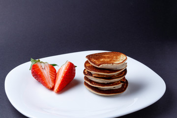 Stack of tasty homemade pancakes laying on white plate with halfes of strawberry on dark background. Top copy space