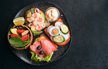 Open sandwiches with salmon, shrimp, radish, cucumber, egg salad, arugula and olives on rye bread and spinach salad with tomatoes on black wooden background. Top view with copy space.