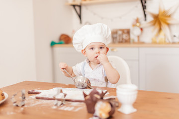Cute little mother helper in cooker hat getting new experience licking a spoon soiled in flour. Infant boy in Chefs Hat playing in kitchen table.